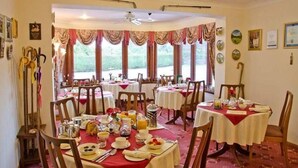 Uma área de refeições com mesas redondas cobertas por toalhas vermelhas, cadeiras de madeira e itens de café da manhã.