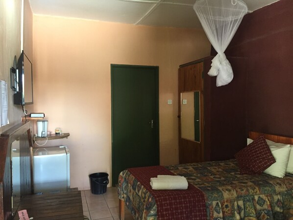 A hotel room with a bed, a bedside table, a mirror, a green door, and a small trash bin. - Alfa Accommodation (Maun)