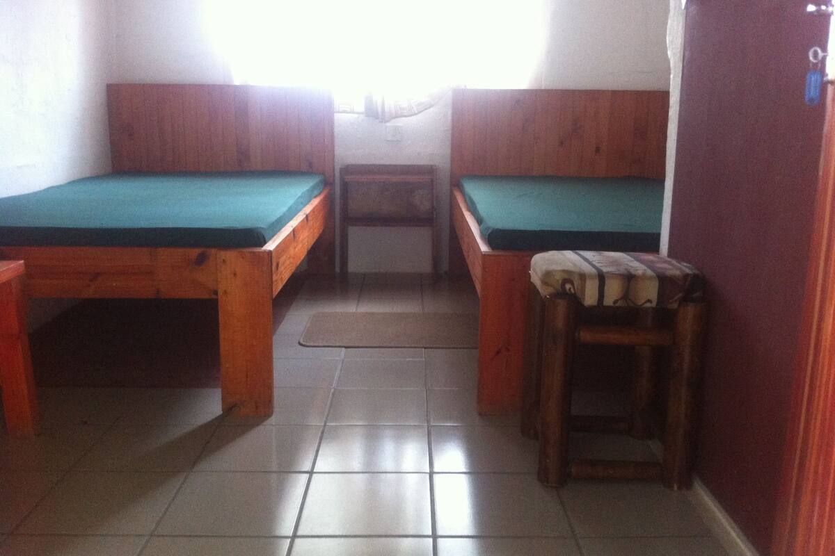 Una habitación pequeña con dos camas de madera con colchones verdes, una mesita de madera y un banco.