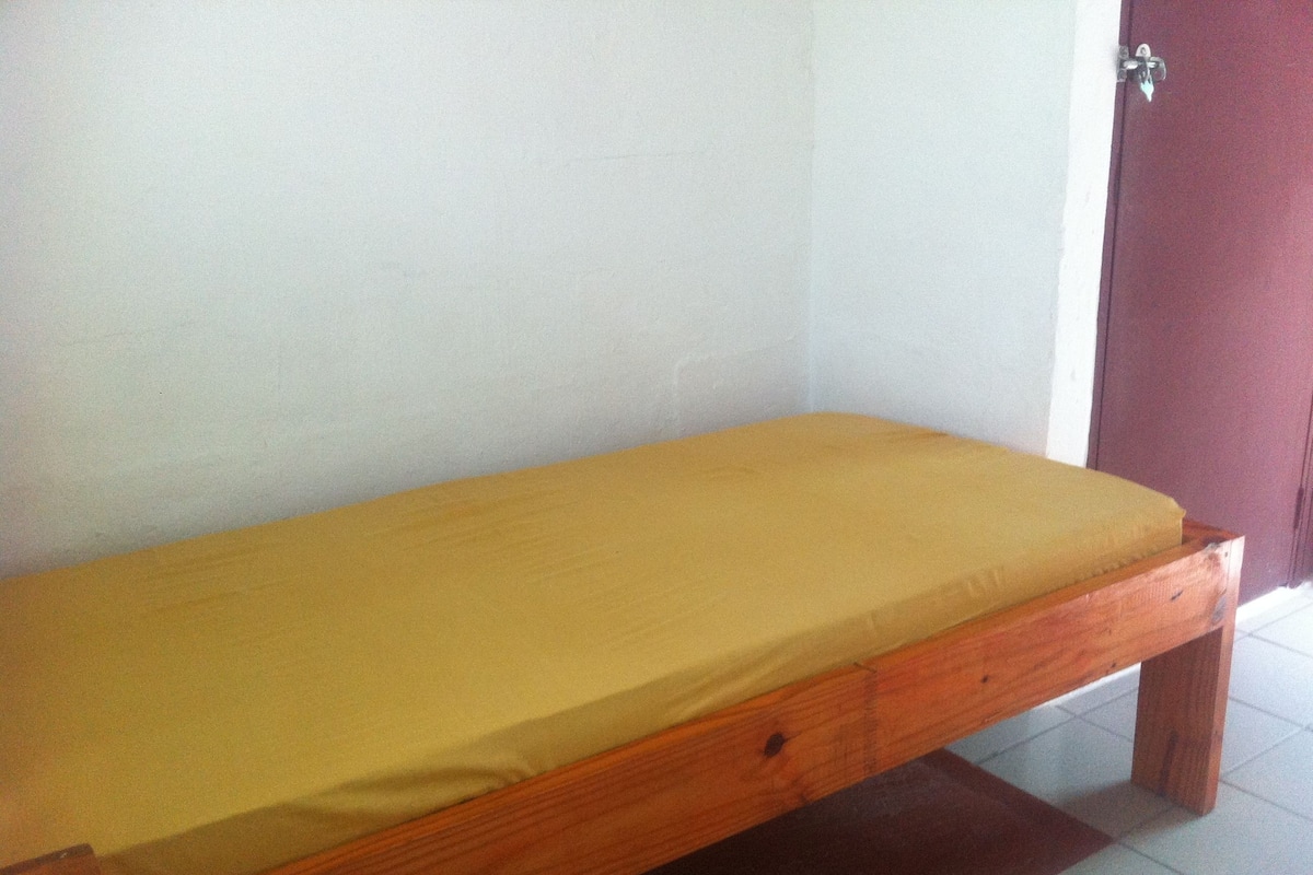 Una cama de madera individual con colchón amarillo en una habitación con paredes blancas y una puerta roja.