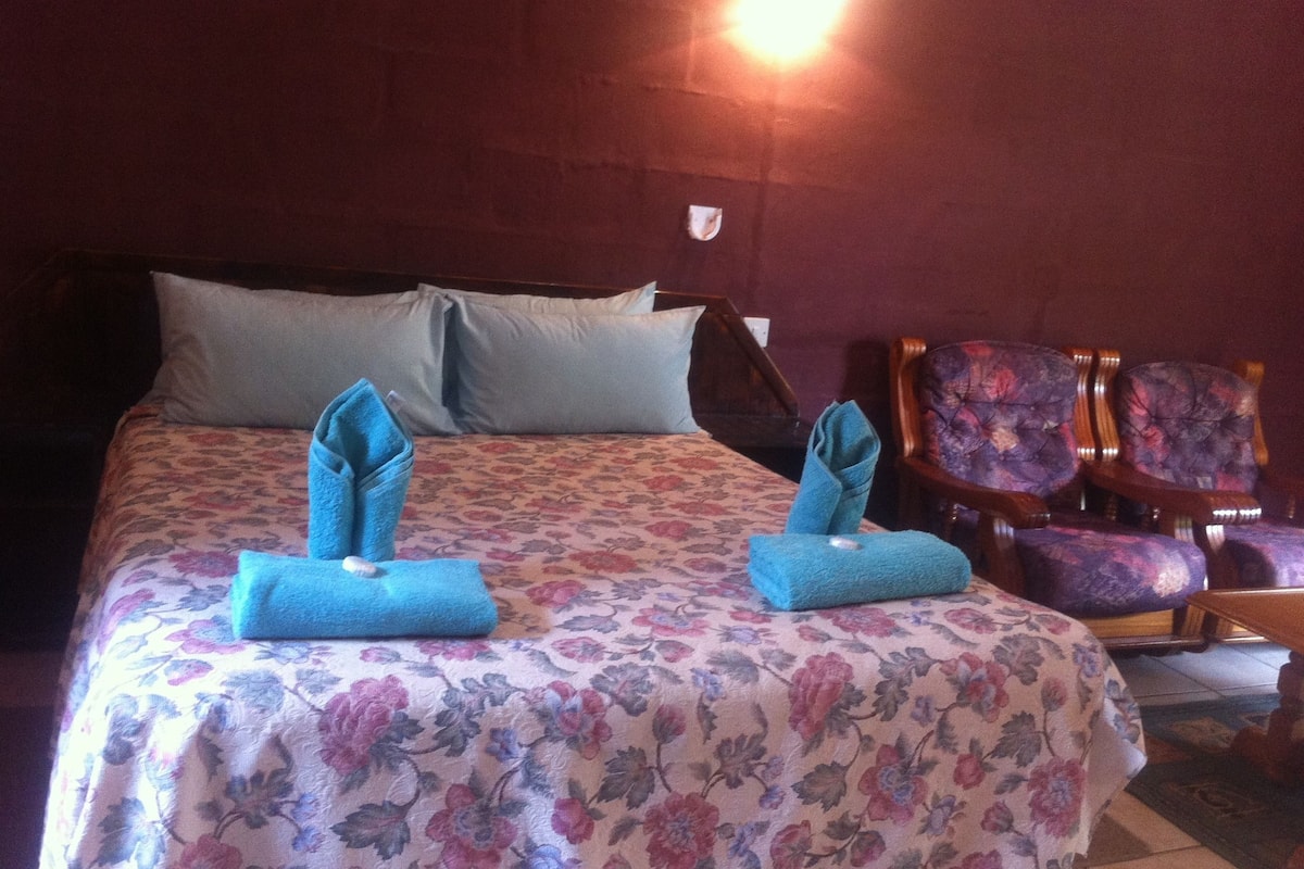 Habitación de hotel con cama, dos toallas dobladas sobre la cama, colcha estampada con motivos florales y una silla con diseño floral.