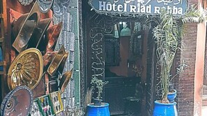 Un escaparate con un letrero que dice "Hotel Rial Rifqa" y diversos artículos expuestos, incluyendo platos de metal, cristalería de colores y plantas en macetas.