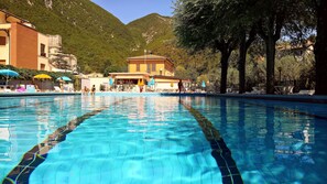A swimming pool with clear blue water, surrounded by trees and buildings. - Hotel Terme San Vittore (Genga)