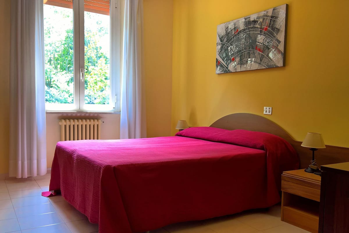 A bedroom with a bed, a radiator, a window with curtains, a nightstand with a lamp, and a wall painting.