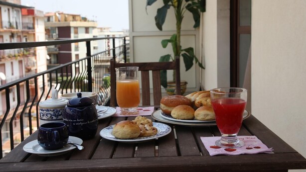 A balcony table with a breakfast setting including a teapot, cups, a glass of orange juice, pastries, and a glass of red drink. - Selene Policlinico di Catania (Gravina di Catania)