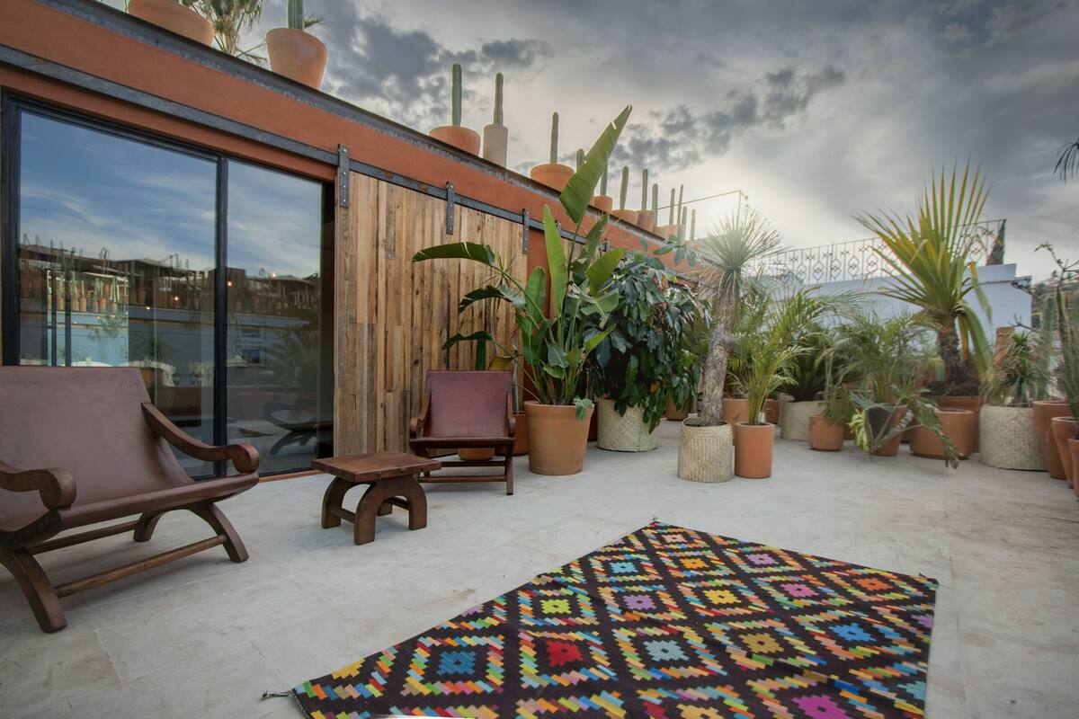 A patio with wooden furniture, a colorful rug, and various potted plants.
