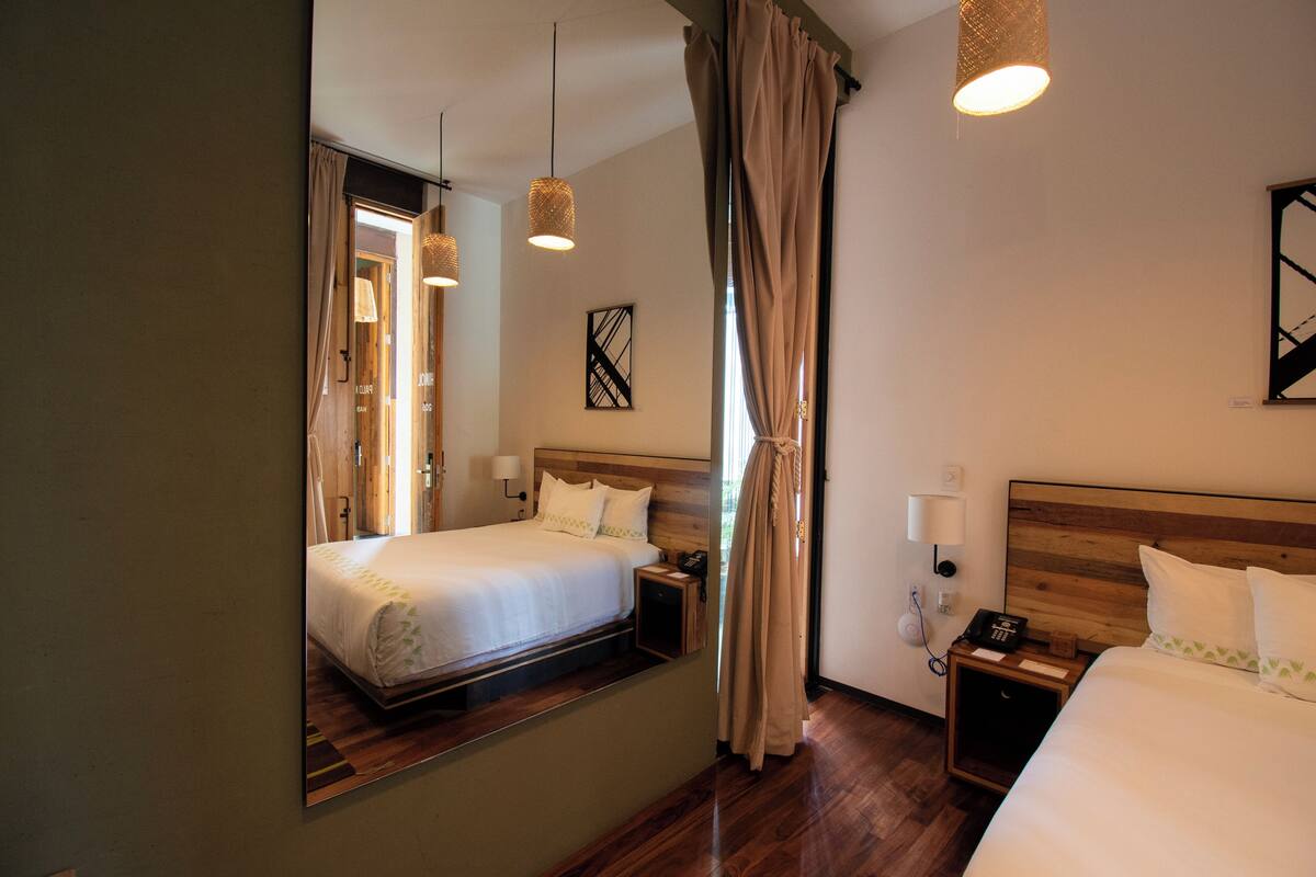 A hotel room with a large mirror reflecting the bed and window, wooden headboard, bedside table with a lamp, and a nightstand with a phone.