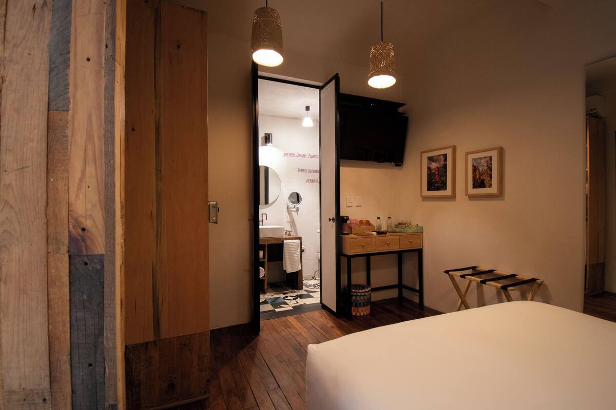 A bedroom with a wooden floor, a bed, a desk with a chair, and a television on the wall.