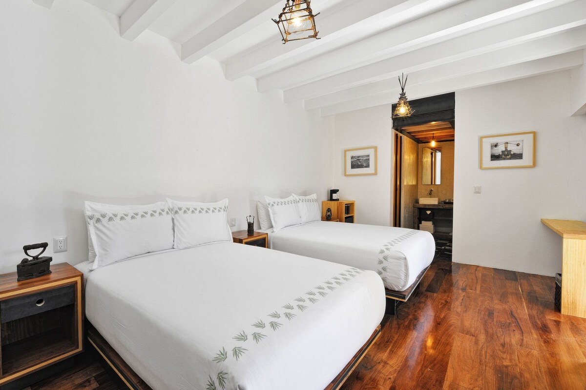 A hotel room with two beds, a wooden floor, a small table, and a bathroom visible through an open doorway.