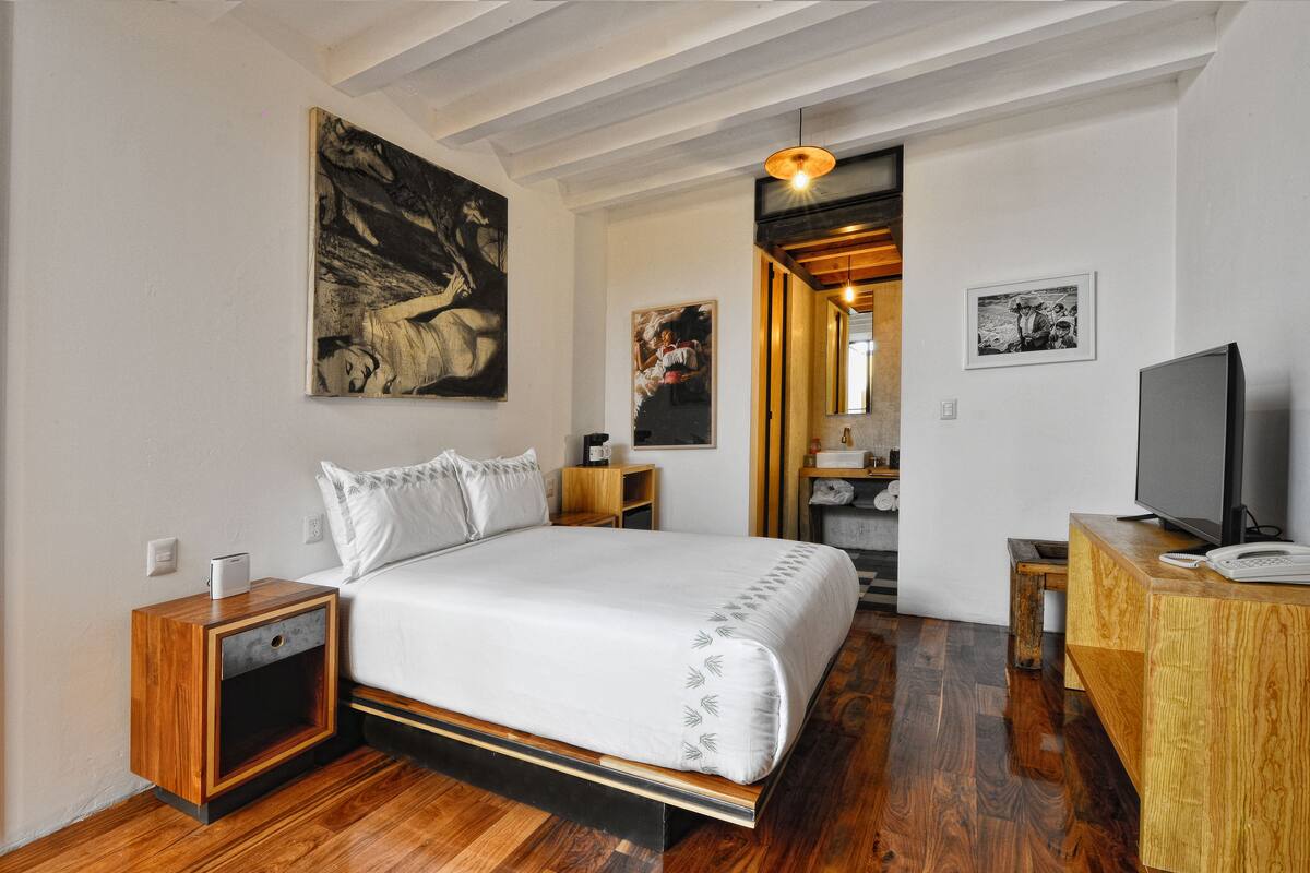 A bedroom with a wooden bed, a nightstand, a desk, and a television. There are paintings on the walls and a wooden ceiling.