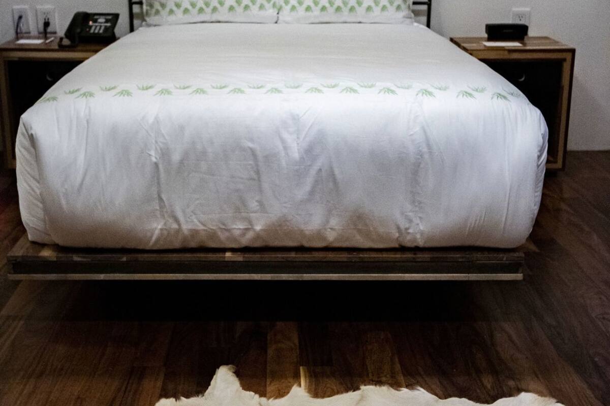 A bed with a white bedspread and green leaf pattern, a wooden headboard, a bedside table with a phone and a lamp, and a fur rug on the floor.