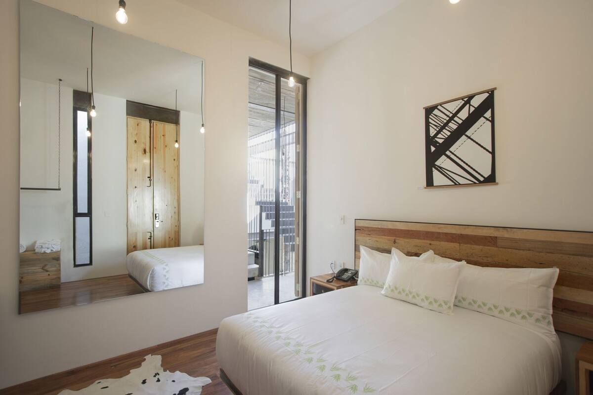 A bedroom with a large bed, a wooden headboard, a mirror, a phone on a bedside table, and a view through a glass door.