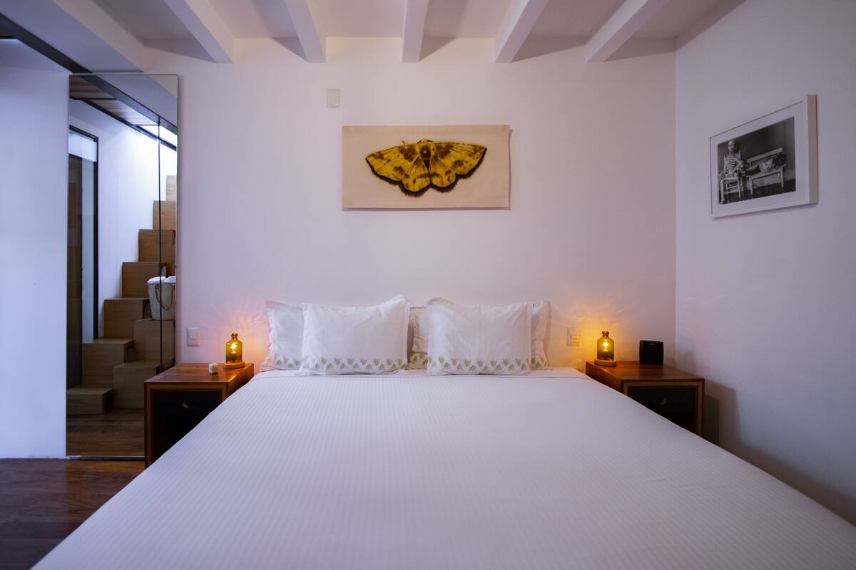 A bedroom with a large bed, two bedside tables with lamps, a framed artwork above the bed, and a staircase leading to an upper floor.