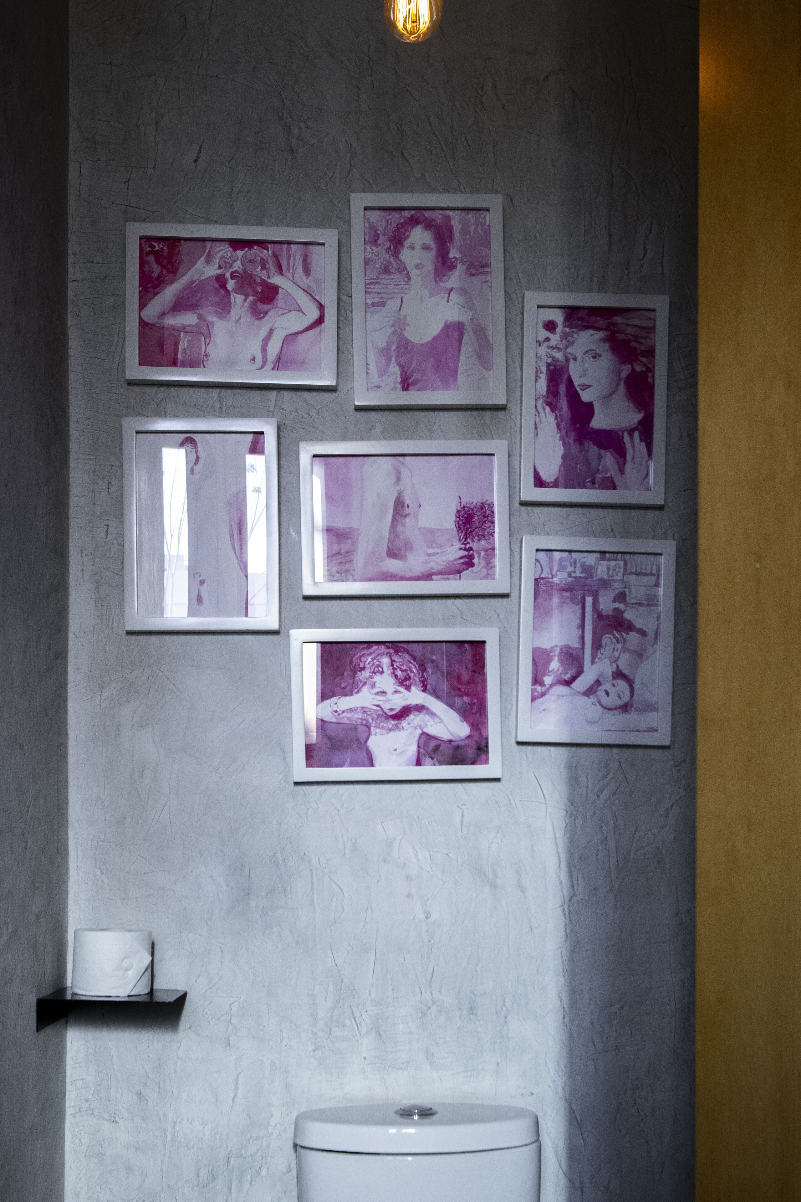 A bathroom with a white toilet, a shelf with a roll of toilet paper, and six framed pictures on the wall.