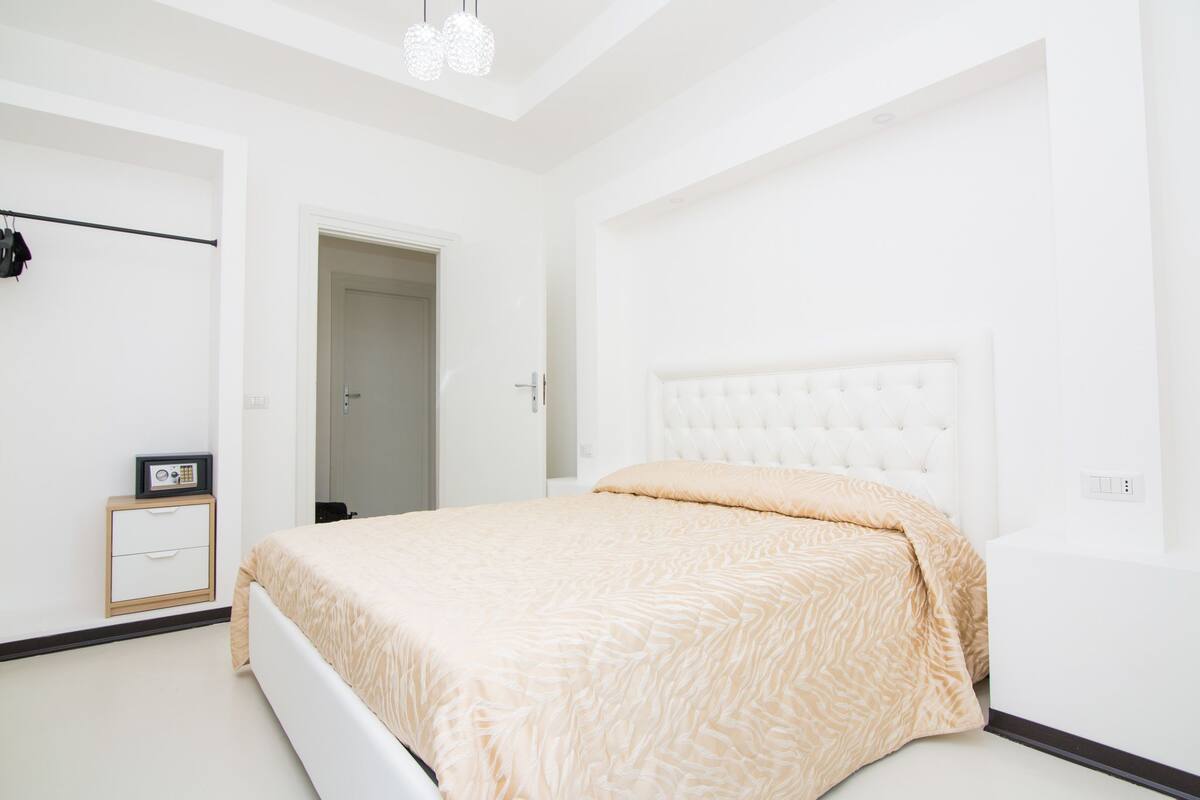 A bedroom with a white bed, a bedside table with a radio, and a wardrobe with clothes hanging.