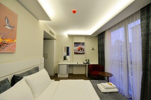 A hotel room with a bed, a desk, a chair, a small table, a mirror, and a window with curtains. - Zeytindali Hotel (Trabzon)