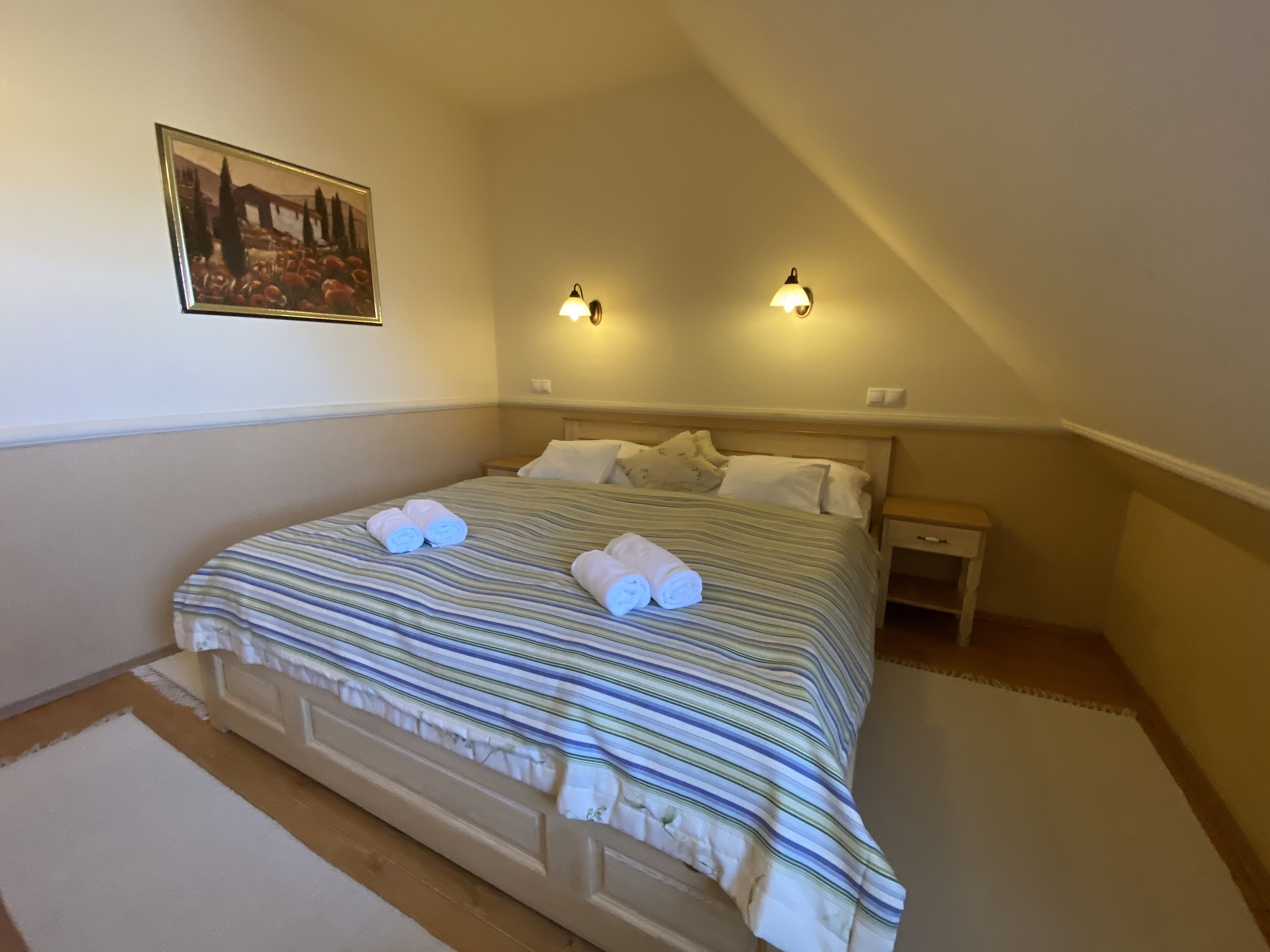Ein ordentlich bezogenes Bett mit gestreifter Bettwäsche und gerollten Handtüchern, ein Nachttisch, eine Wandlampe.