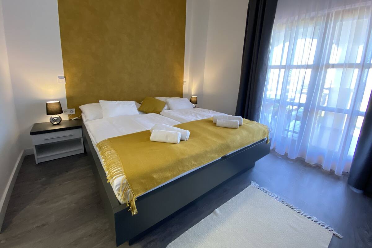 Habitación de hotel con una cama grande, dos mesitas de noche con lámparas, una pared decorada con detalles dorados y vistas a través de cortinas translúcidas.