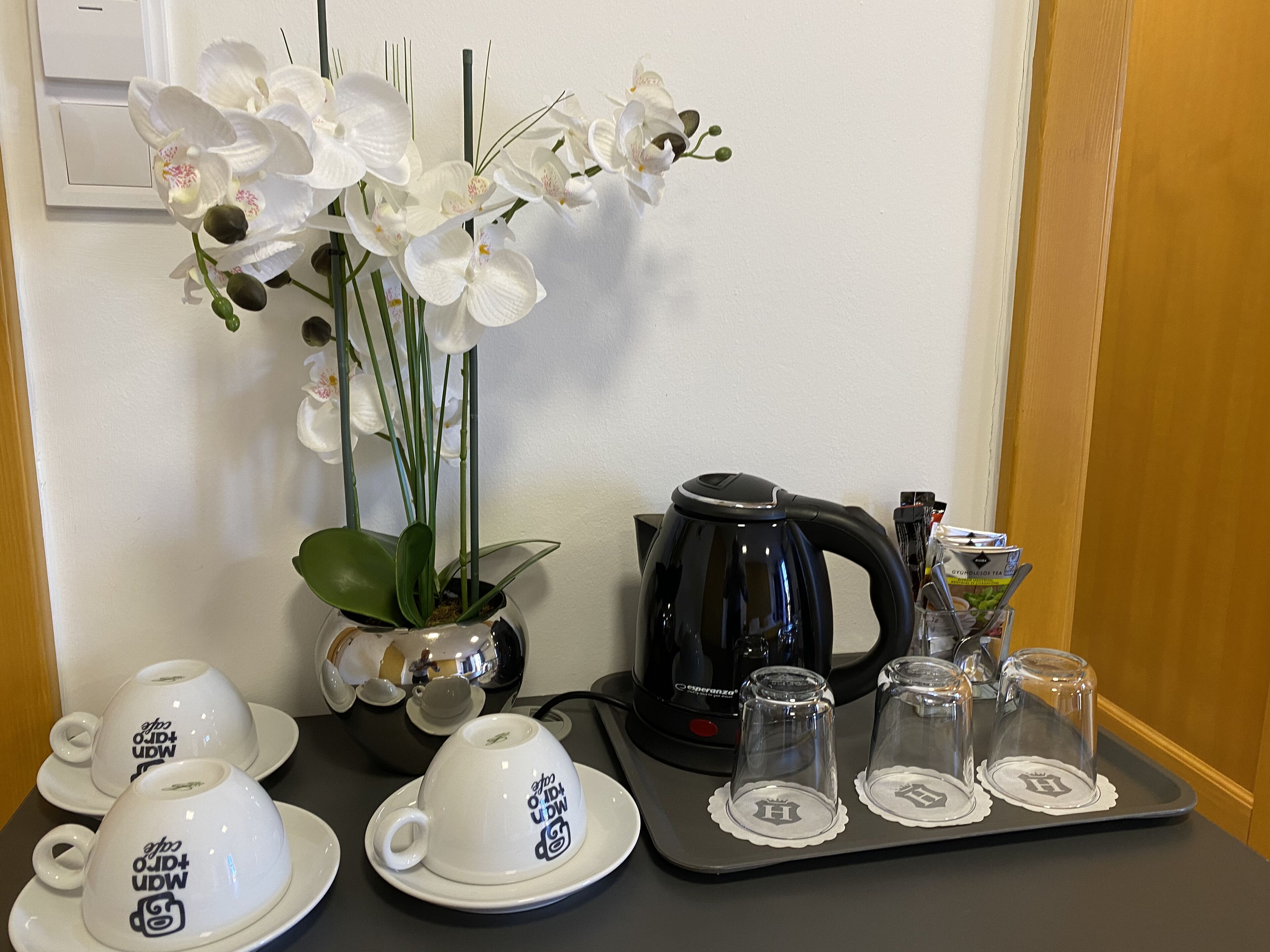 Uma mesa com chávenas brancas, uma chaleira preta, uma planta em vaso e um tabuleiro com copos de vidro.