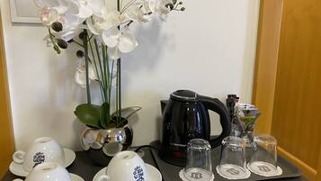 Uma mesa com chávenas brancas, uma chaleira preta, uma planta em vaso e um tabuleiro com copos de vidro.
