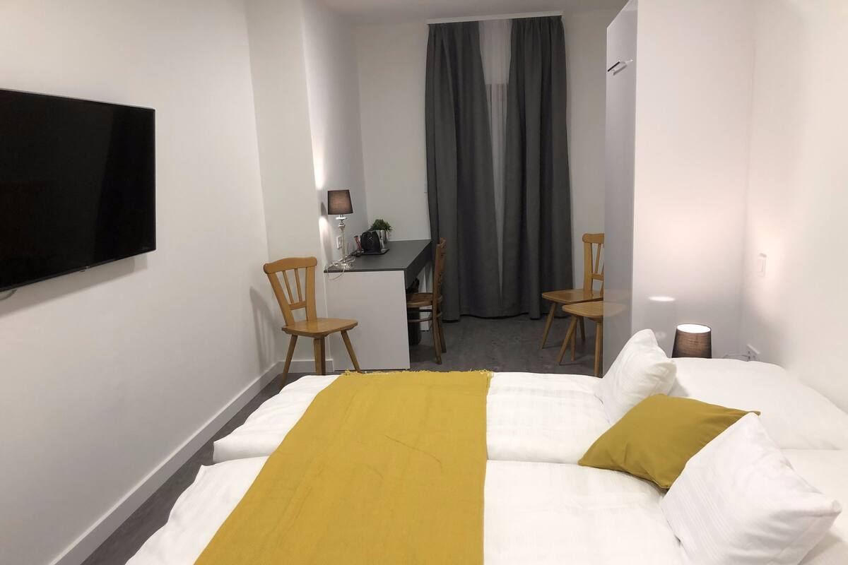 Una habitación de hotel con cama, escritorio, dos sillas, televisor y ventana con cortinas.