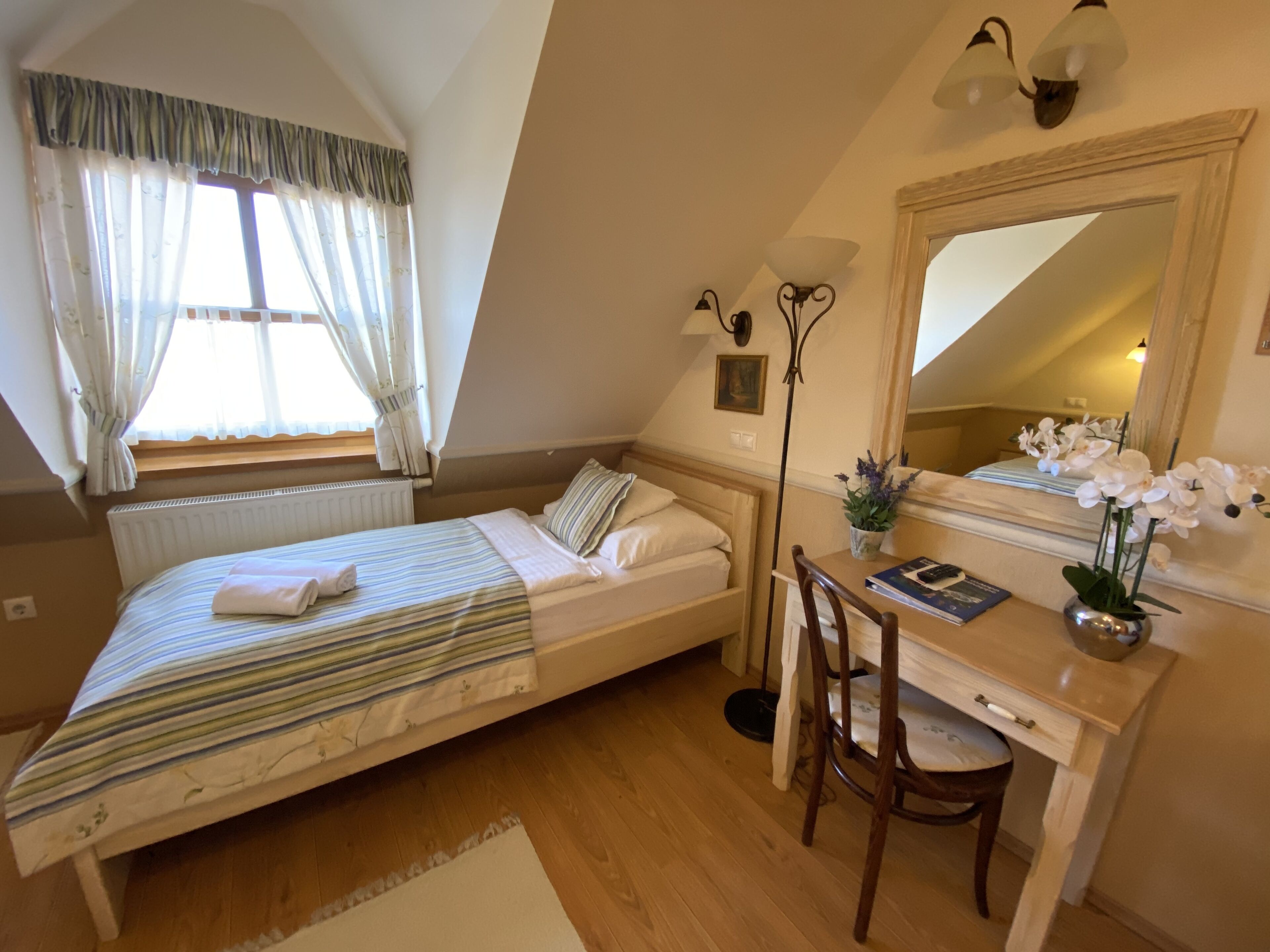 Ein Schlafzimmer mit Bett, Schreibtisch, Stuhl, Heizkörper, einem Fenster mit Vorhängen und einem Spiegel.