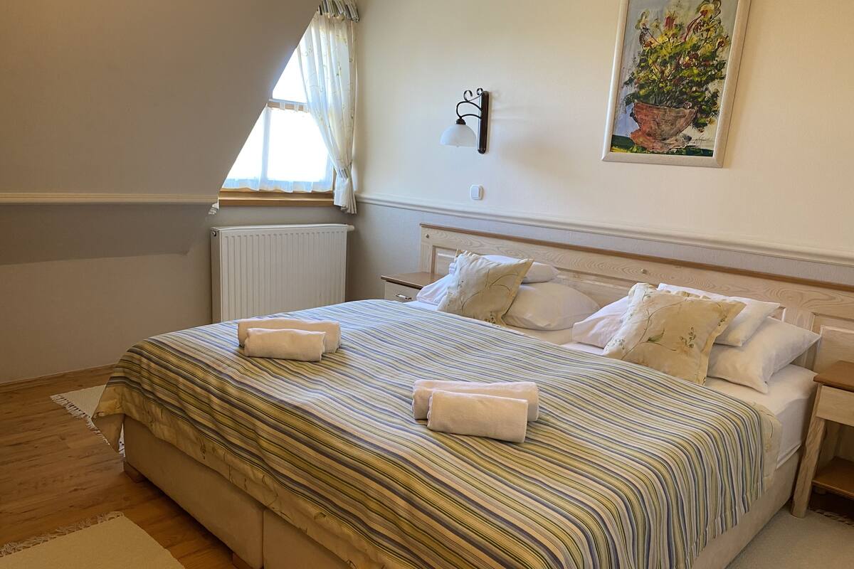 Una cama bien hecha con ropa de cama a rayas y dos toallas, una mesita de noche con una lámpara, un radiador y una ventana con cortinas.