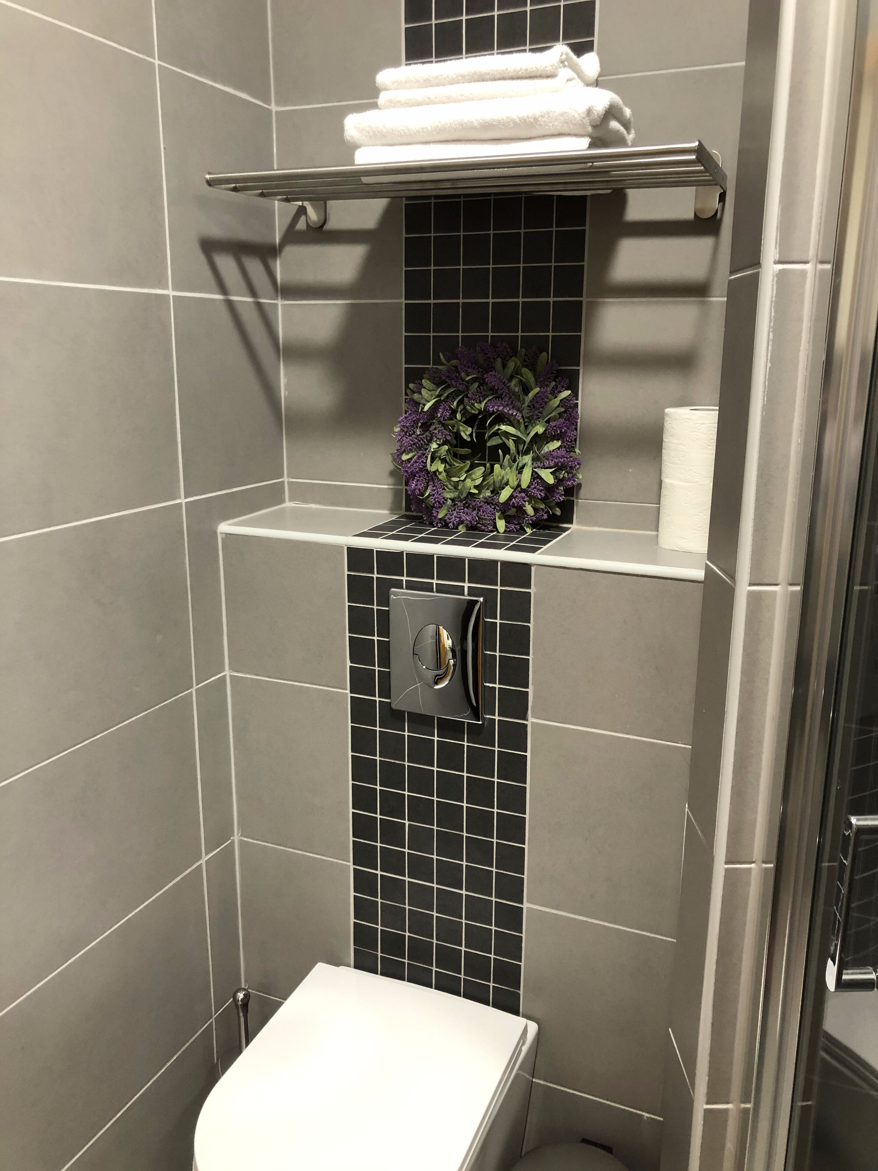 A bathroom with a toilet, a shelf with towels, a wall-mounted toilet paper holder, and a decorative purple wreath.