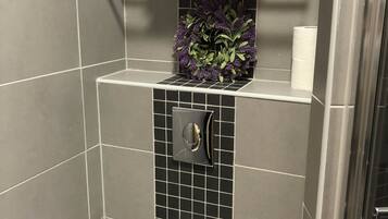 A bathroom with a toilet, a shelf with towels, a wall-mounted toilet paper holder, and a decorative purple wreath.