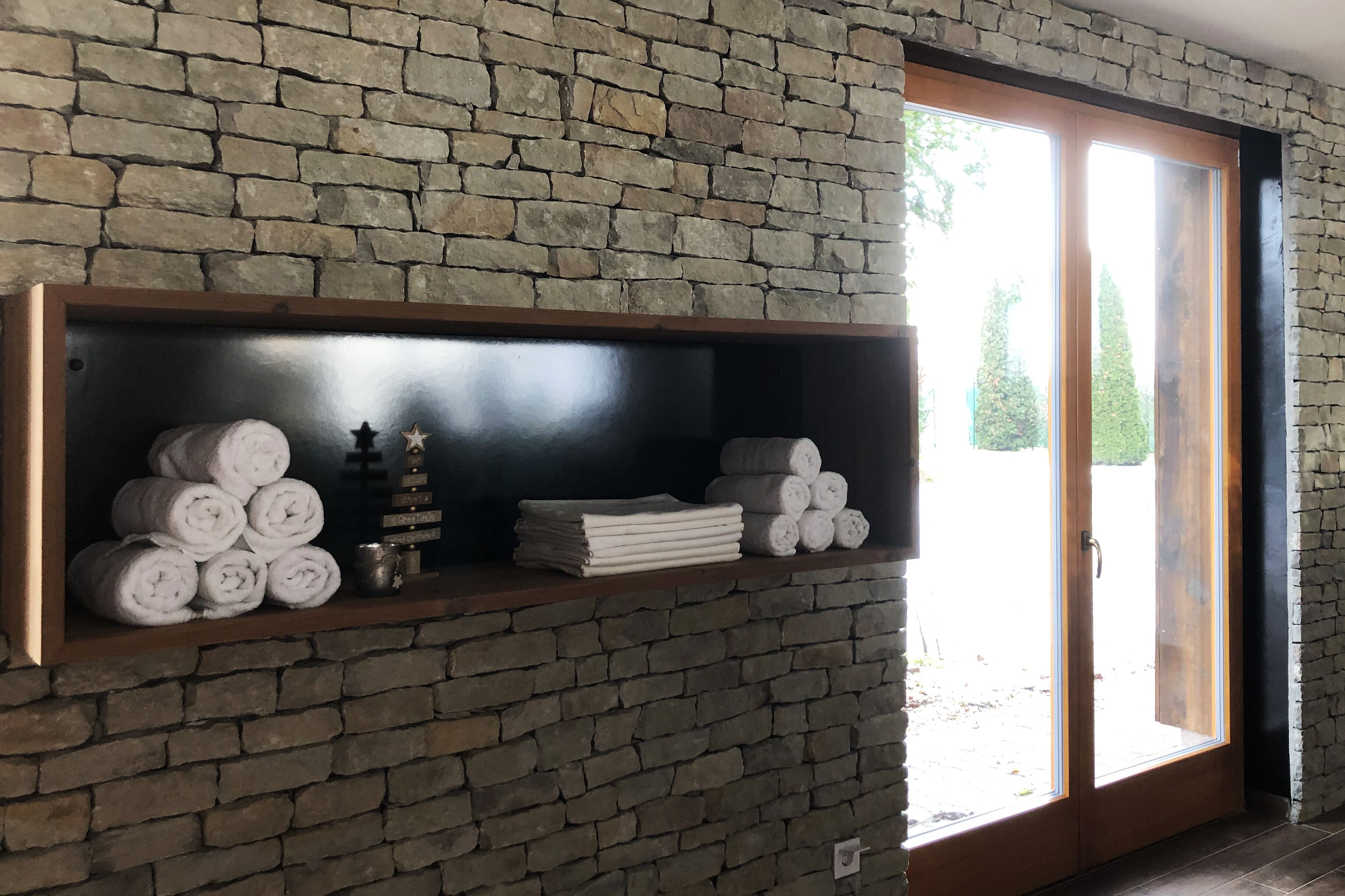 A room with a brick wall, a wooden shelf with rolled towels and a small decorative item, and a sliding glass door leading outside.