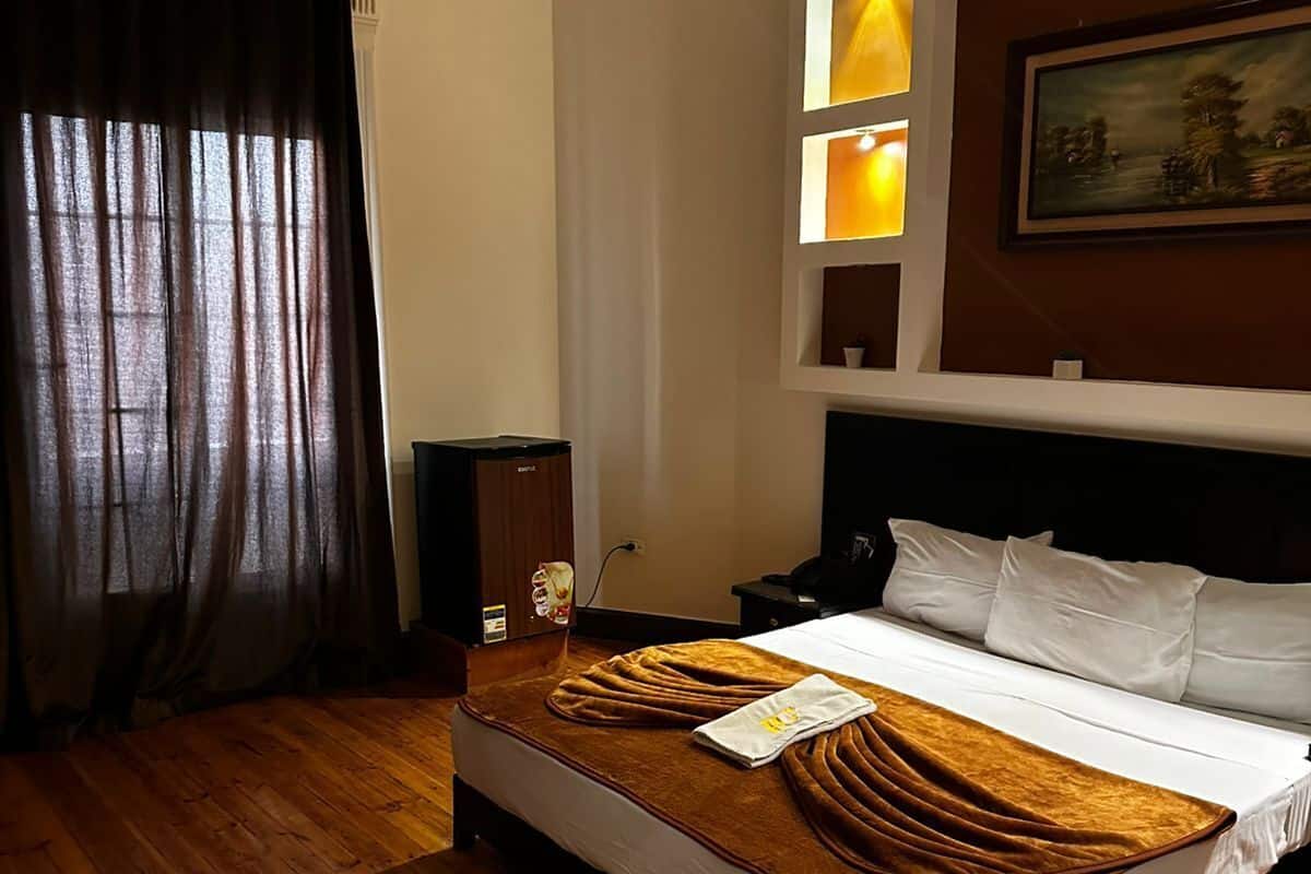 A hotel room with a wooden floor, a bed with white bedding, a brown throw blanket, a bedside table, a lamp, and a painting on the wall.