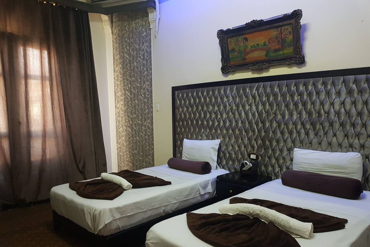 A hotel room with two beds, a large padded headboard, a painting on the wall, and a window with curtains.
