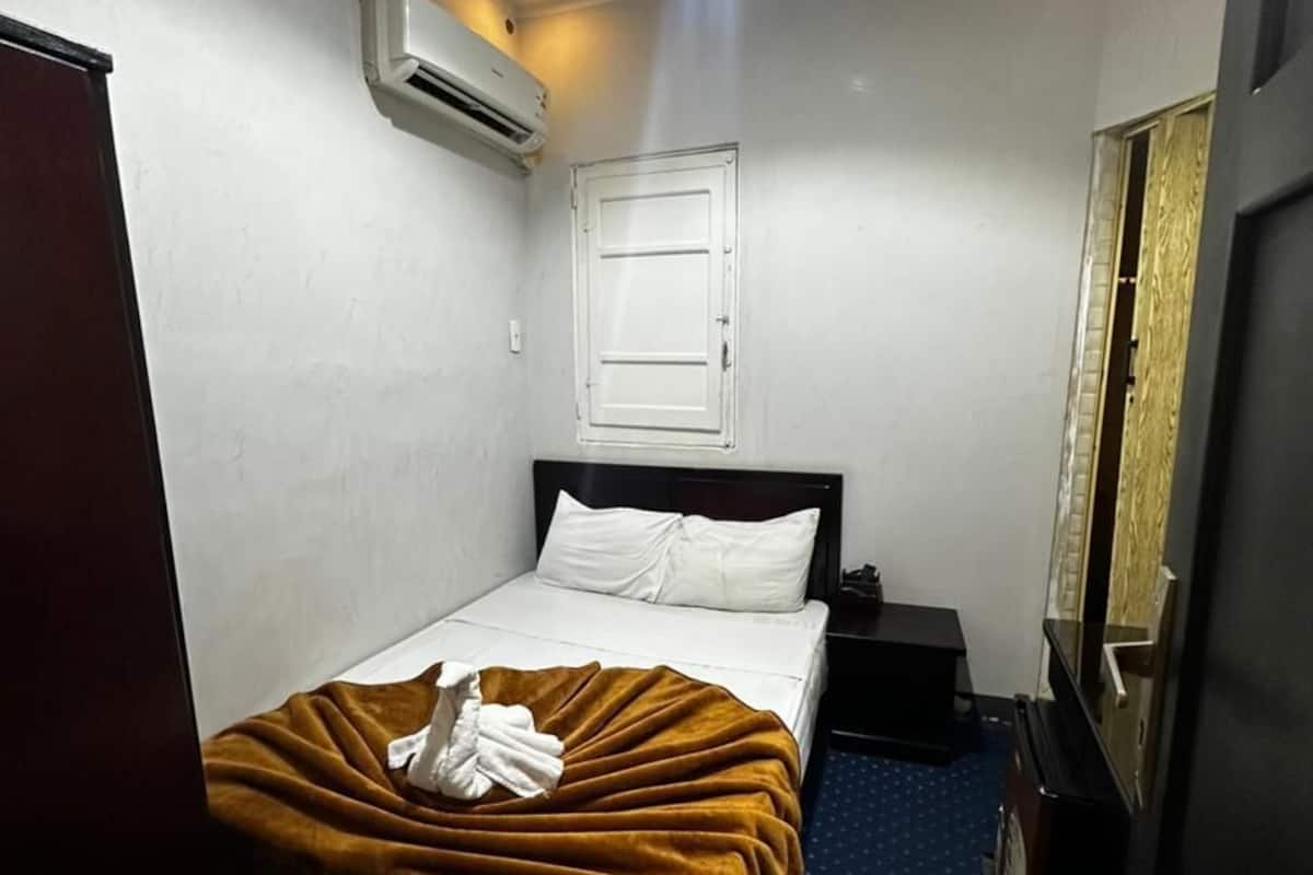 A hotel room with a bed, a nightstand, a wall-mounted air conditioner, and a window with a closed blind.