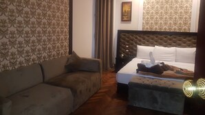 Ein Hotelzimmer mit einer Couch, einem Bett mit einer gepolsterten Kopfteil, einem Nachttisch und einer Wand mit ornamentalem Wandpapier.