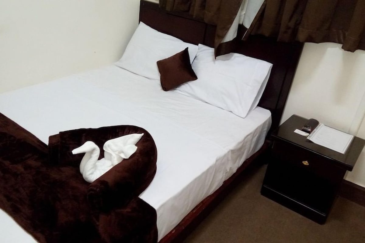 A neatly made bed with white linens and brown pillows, a dark brown throw blanket, a small bedside table with a lamp, and a nightstand with a phone and a notepad.