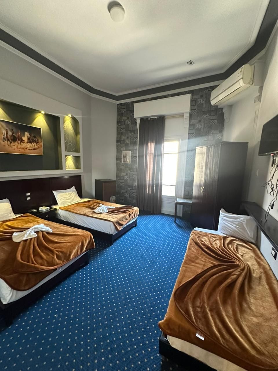 A hotel room with two beds, a large window with curtains, a flat-screen TV, and a wall-mounted air conditioning unit.