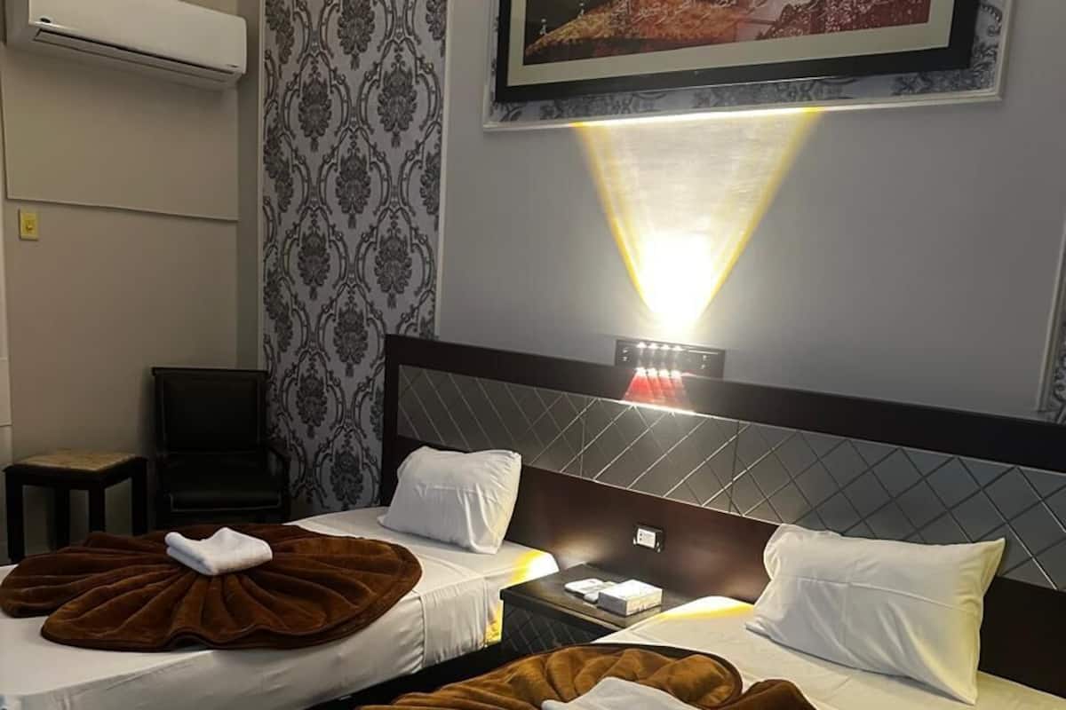 A hotel room with two beds, a wall-mounted air conditioner, a framed picture, and a bedside lamp.