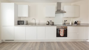 Une cuisine moderne avec des meubles blancs, un four à micro-ondes en acier inoxydable, une bouilloire, un grille-pain et un four dont la porte est ornée d’un torchon.