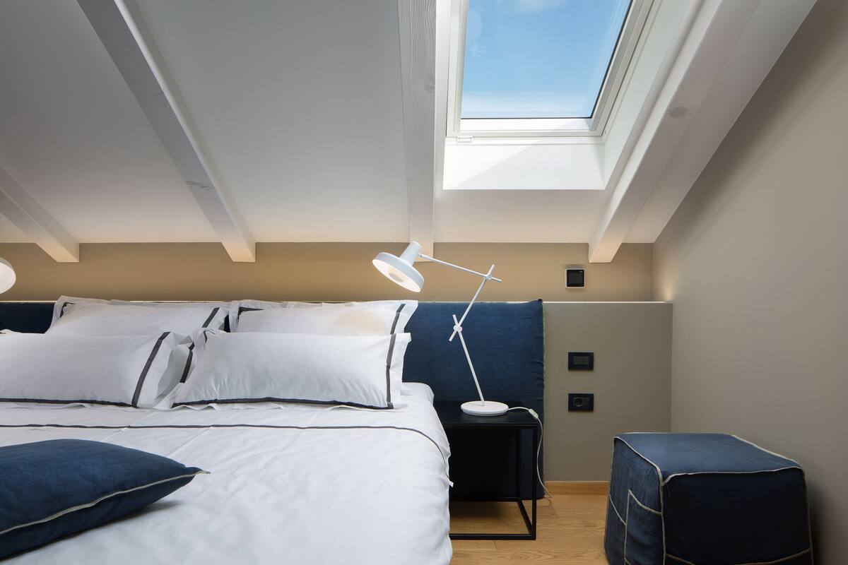 Une chambre avec un puits de lumière, un lit avec une literie blanche, une table de chevet avec une lampe et un petit pouf bleu.