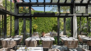 A covered outdoor dining area with tables and chairs, surrounded by glass walls and a view of greenery. - Almayer Art & Heritage Adults Only Hotel & Dépendance Rooms (Zadar)
