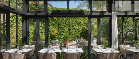 A covered outdoor dining area with tables and chairs, surrounded by glass walls and a view of greenery.