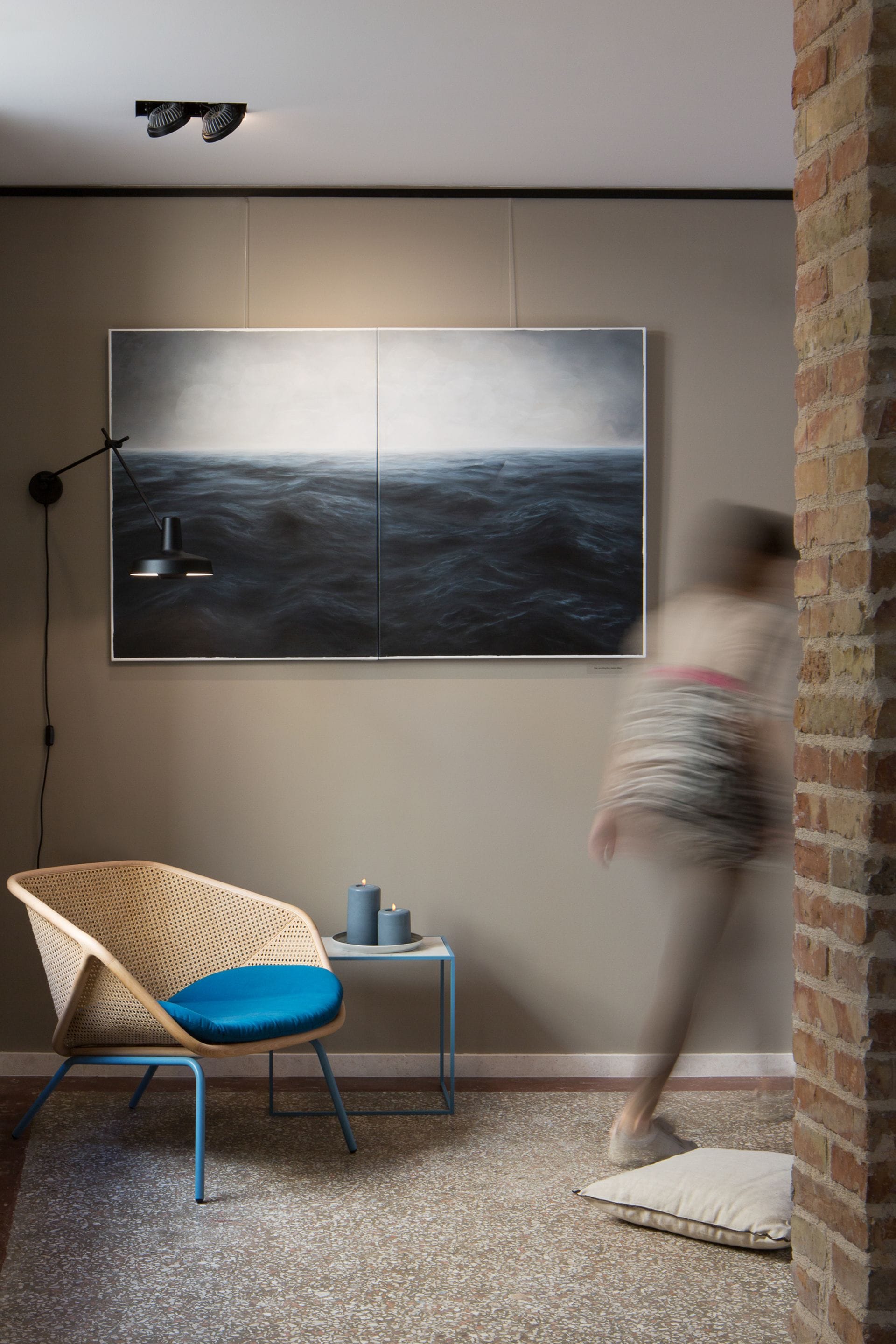 Una habitación con un cuadro abstracto grande, una silla de mimbre con un cojín azul, una mesita con una vela y una pared de ladrillo.