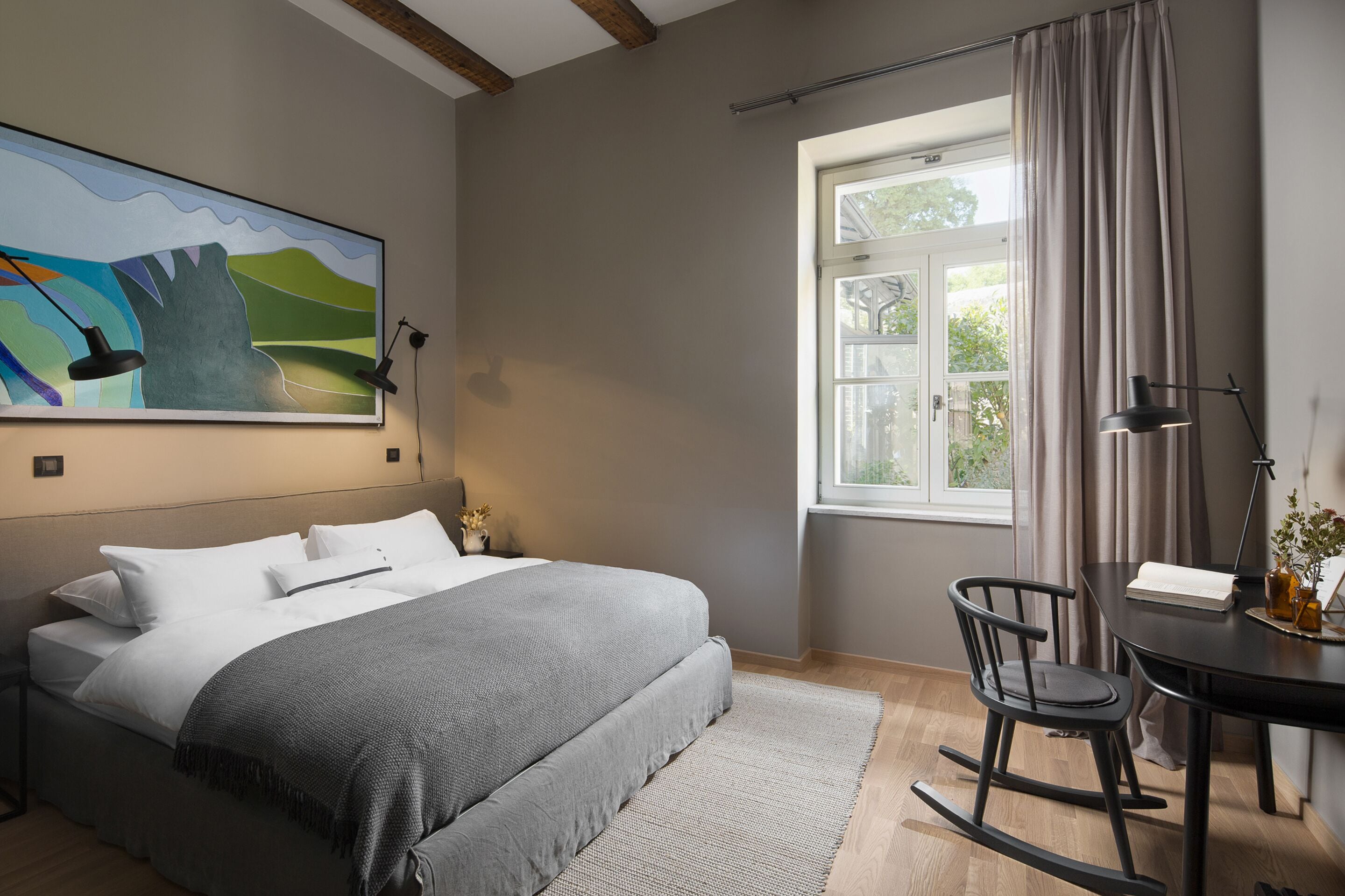 A bedroom with a bed, a desk, a rocking chair, a window with curtains, and a painting on the wall.