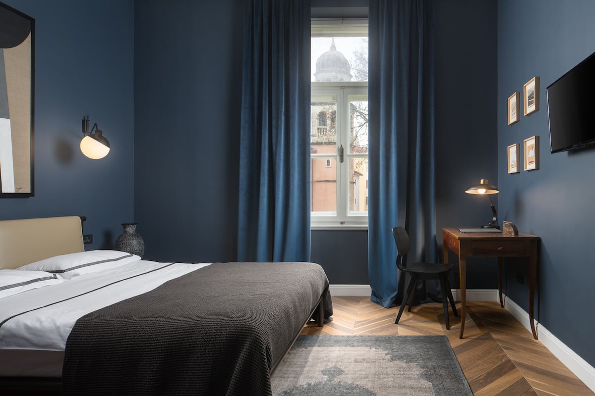 Une chambre à coucher comprenant un lit, un bureau, une chaise, une fenêtre avec des rideaux, un téléviseur fixé au mur et des tableaux encadrés.