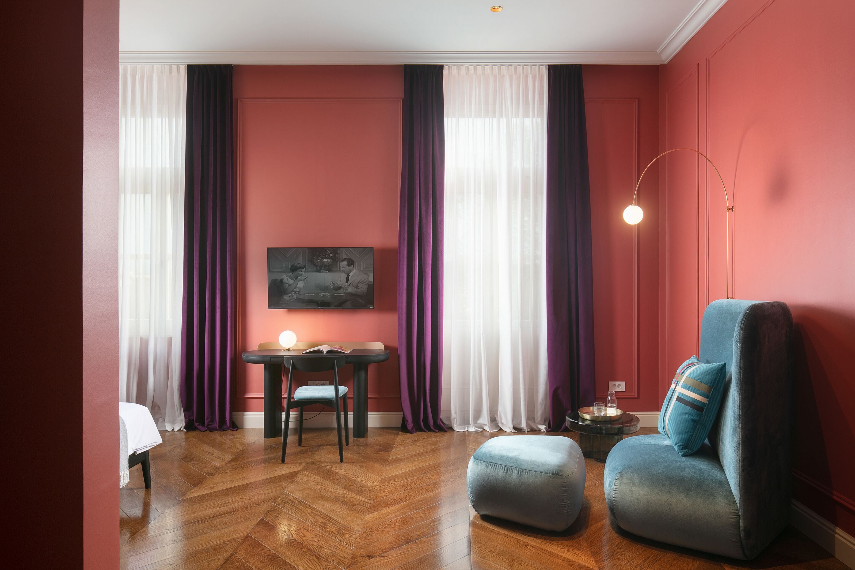 A room with a wooden herringbone floor, a blue armchair, a small ottoman, a round side table with a lamp, a dining table with chairs, and a painting on the wall.
