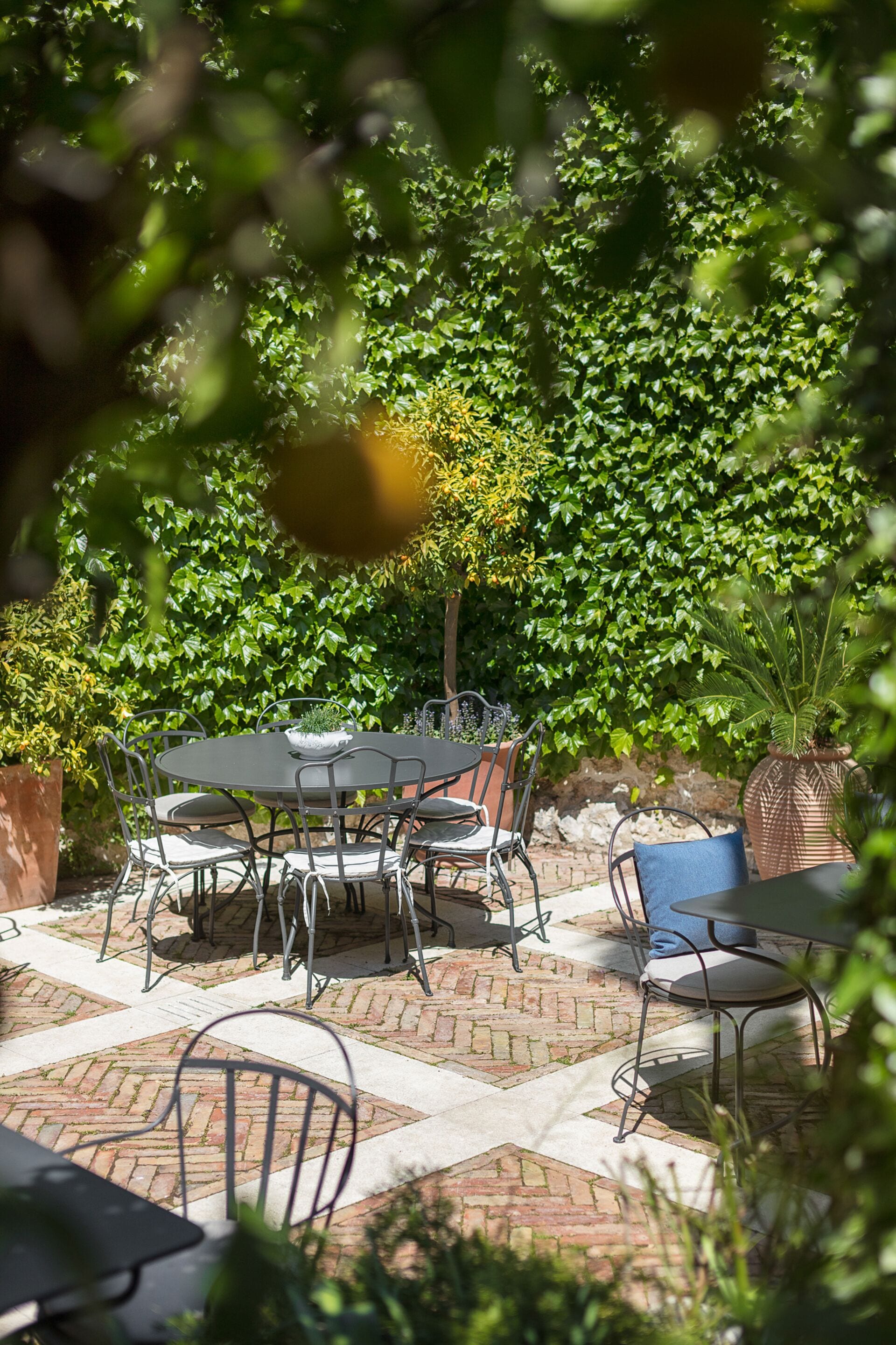Une terrasse extérieure avec un sol en briques, des tables et des chaises en métal, et un décor luxuriant de verdure.