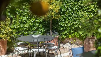 Un patio al aire libre con piso de ladrillo, mesas y sillas de metal, y un exuberante fondo verde.