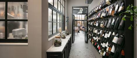Une cave à vin avec une longue rangée de bouteilles de vin alignées sur des étagères, un comptoir sur lequel sont posés une bougie et un vase, et une vue sur l’extérieur par une fenêtre.