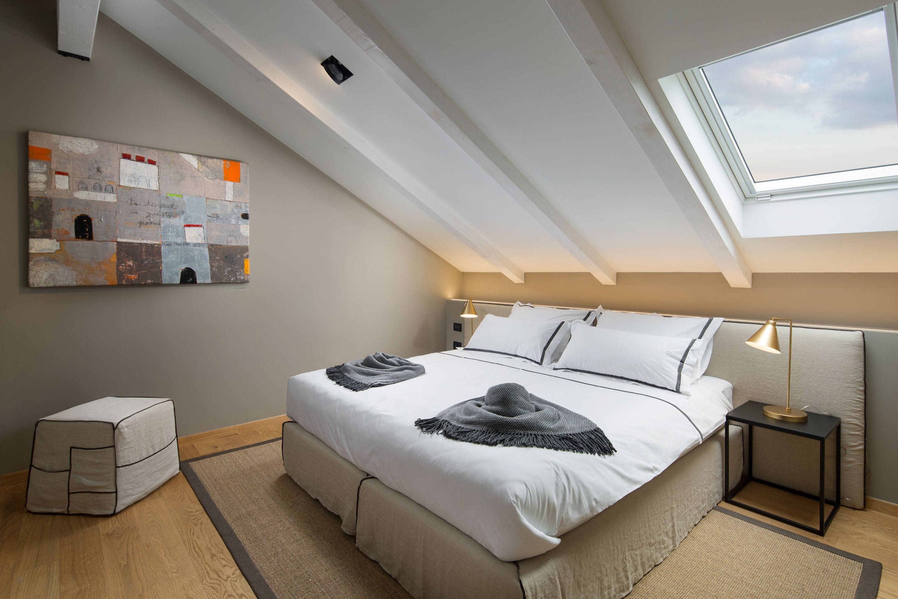 A bedroom with a skylight, a bed with white linens, a bedside table with a lamp, and a wall art piece.