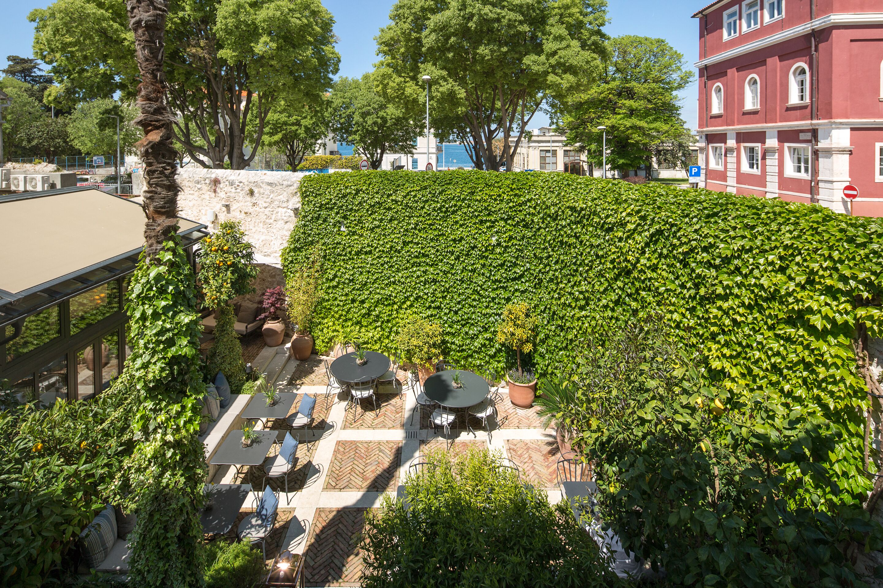 A rooftop patio with tables and chairs, surrounded by lush greenery and a stone wall.